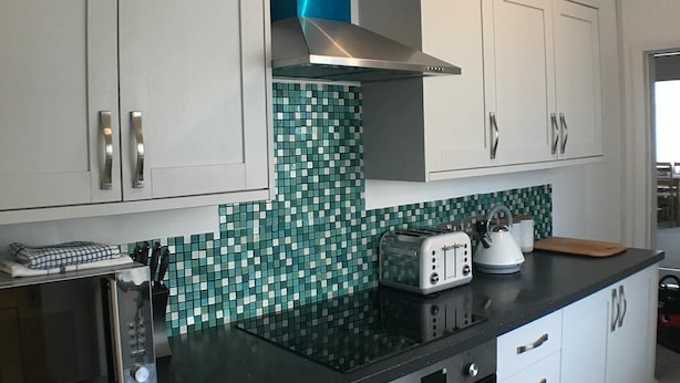 A modern kitchen with white cabinets, a black countertop, a blue range hood, a toaster, and a microwave. - Eastville Court (Rhyl)