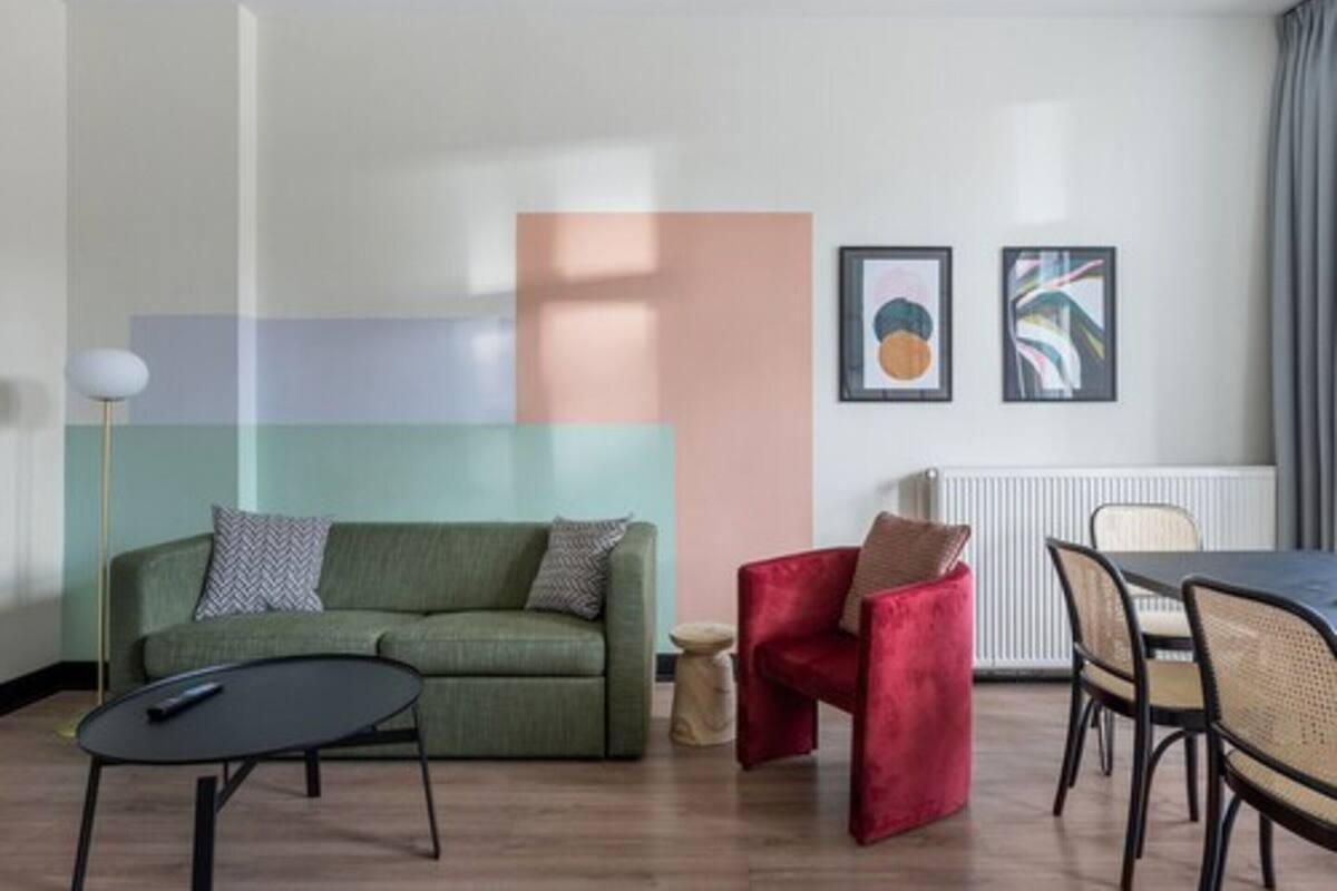 A modern living room with a green sofa, a red chair, a round table, and a dining area with chairs. There are two framed artworks on the wall.
