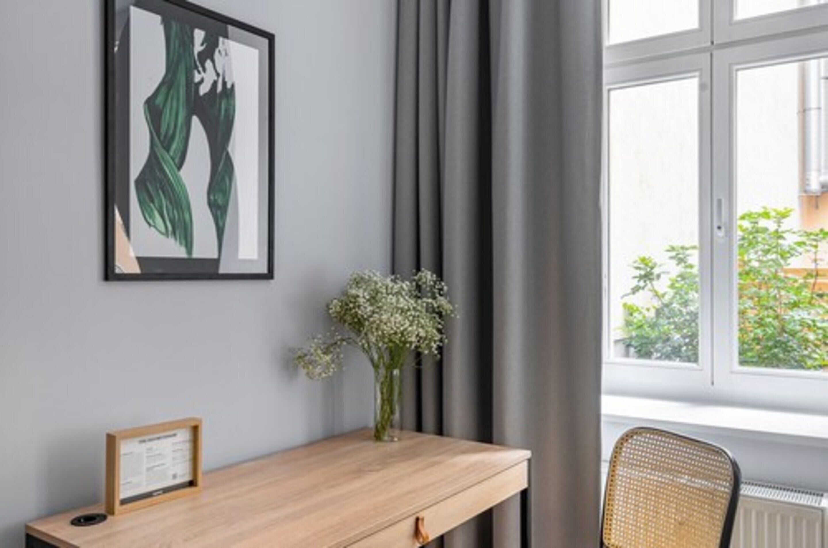 Una habitación con un escritorio de madera, un cuadro enmarcado, un jarrón con flores y una ventana con cortinas.