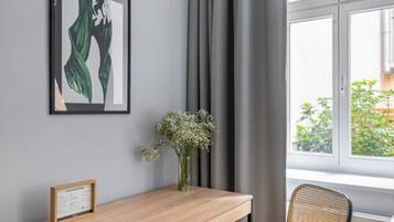 Een kamer met een houten bureau, een lijst met een afbeelding, een vaas met bloemen en een raam met gordijnen.