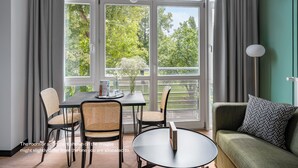 Moderni huone, jossa on ruokapöytä, tuoleja ja sohva. Suuret ikkunat, joissa on verhot, tarjoavat näkymän ulkona olevaan vehreään maisemaan.