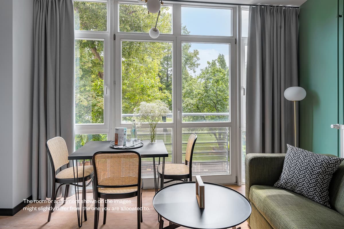 A modern room with a dining table, chairs, and a sofa. Large windows with curtains provide a view of greenery outside.