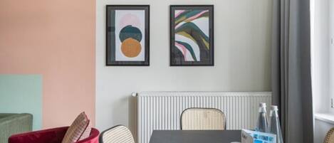 Une chambre d’hôtel moderne avec une table à manger, deux tableaux abstraits encadrés, un radiateur et une fenêtre avec des rideaux.