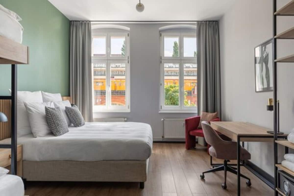 A modern hotel room with a large bed, a desk, and a view of the city through large windows.