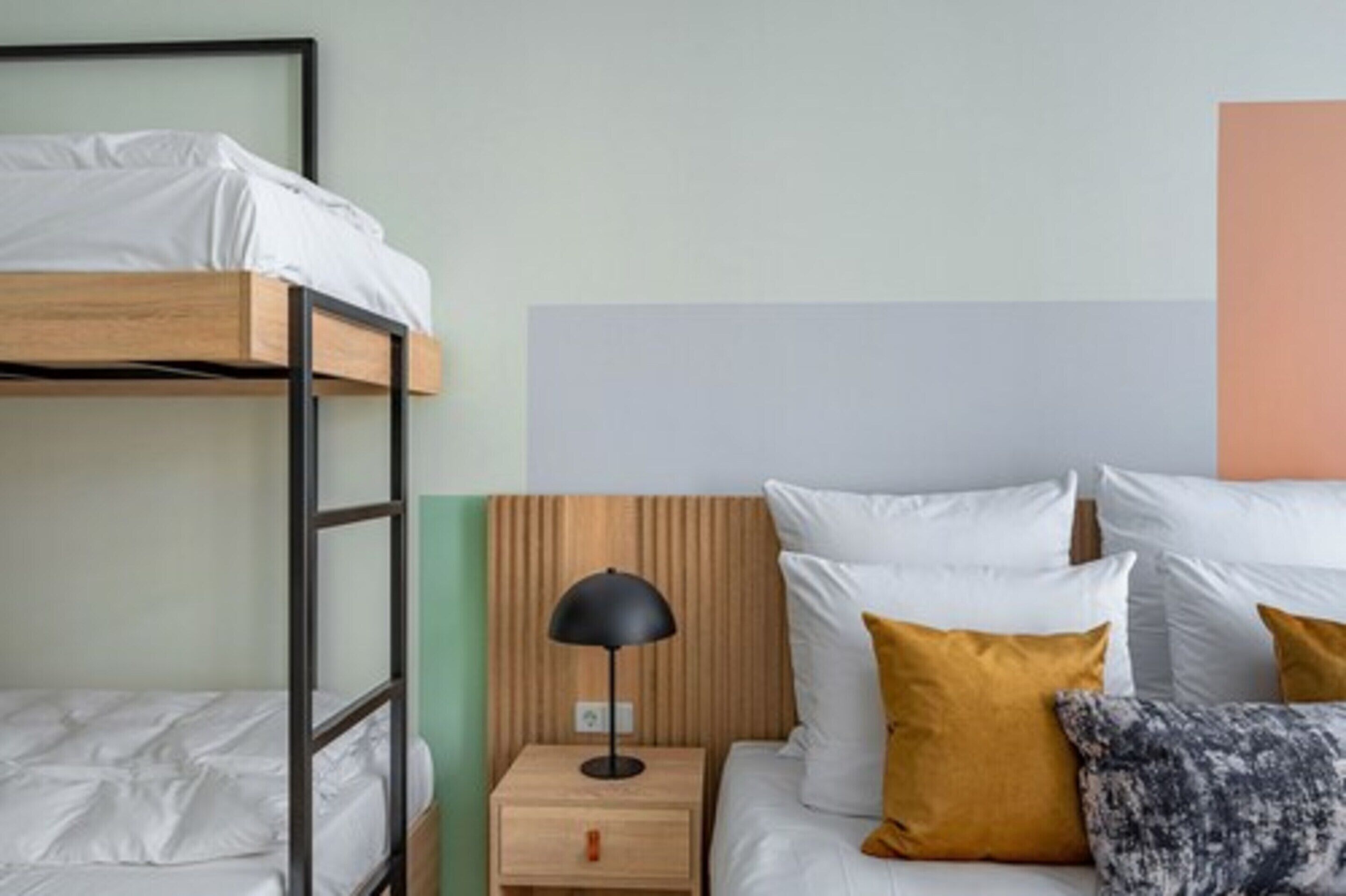 Una cama individual en literas con estructura de madera, una mesita de noche con lámpara, y otra cama con ropa de cama blanca y dos almohadas.