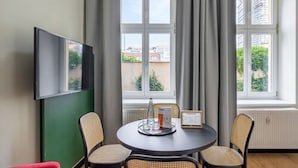 Hotellihuone, jossa on pyöreä pöytä, kaksi rottinkituolia, seinälle asennettu litteänäyttöinen televisio ja ikkuna, jossa on verhot.