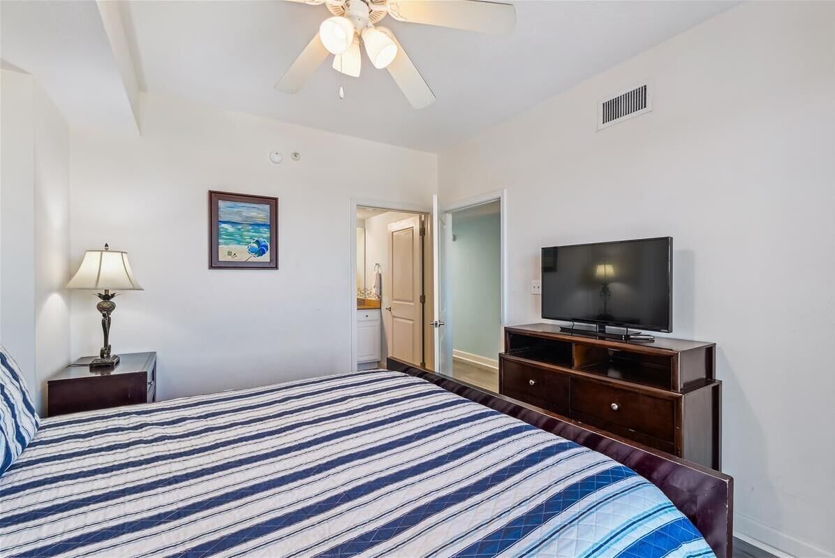 A hotel room with a bed, a television, a ceiling fan, a lamp, and a painting on the wall.