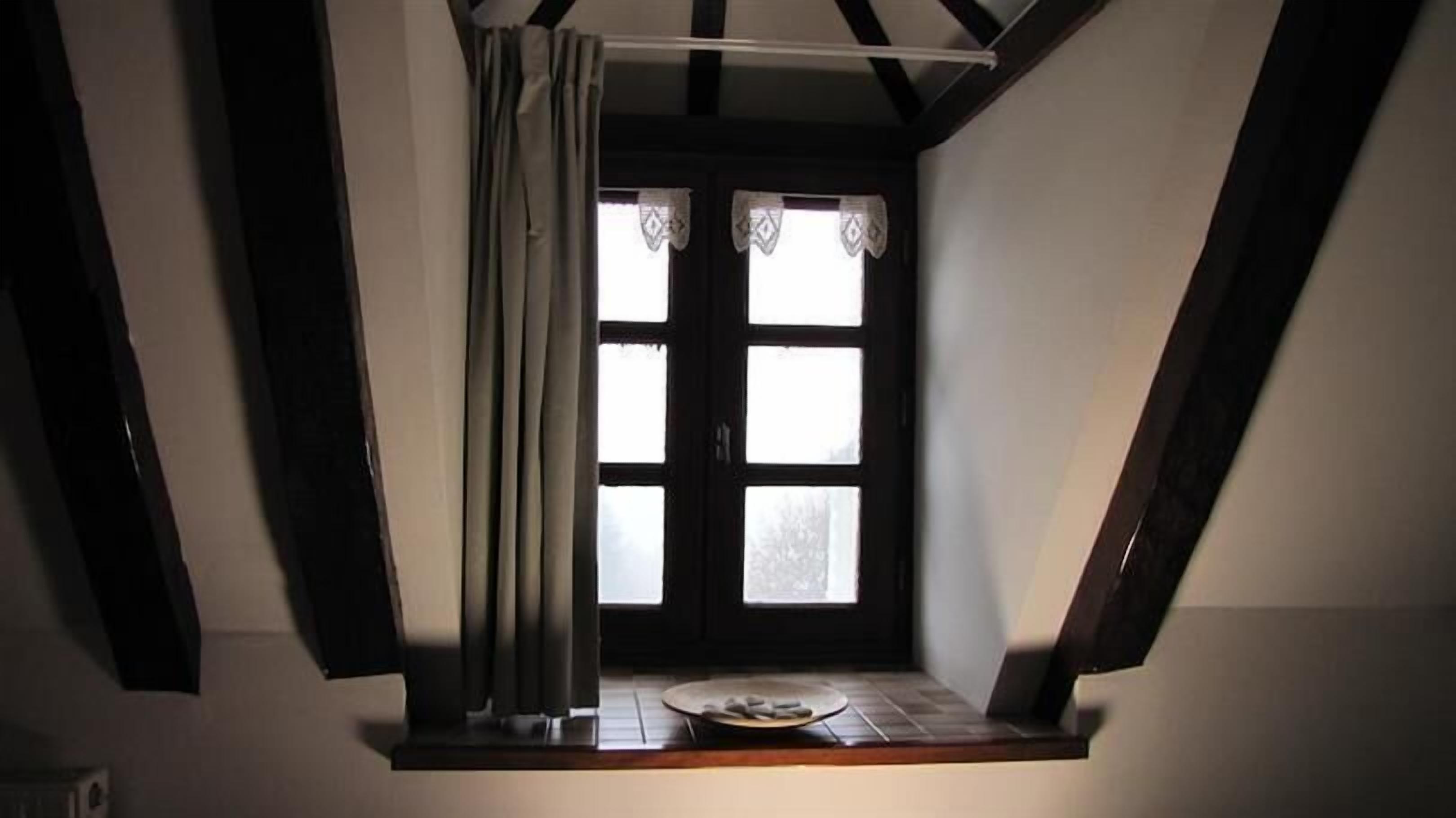 Une pièce avec un plafond en pente, une fenêtre avec des rideaux et une petite table sur laquelle se trouve une assiette.