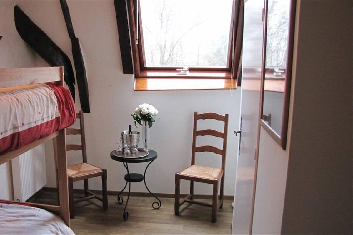 Een kleine kamer met een stapelbed, twee houten stoelen, een tafeltje met een bloevas en een raam met uitzicht op bomen.