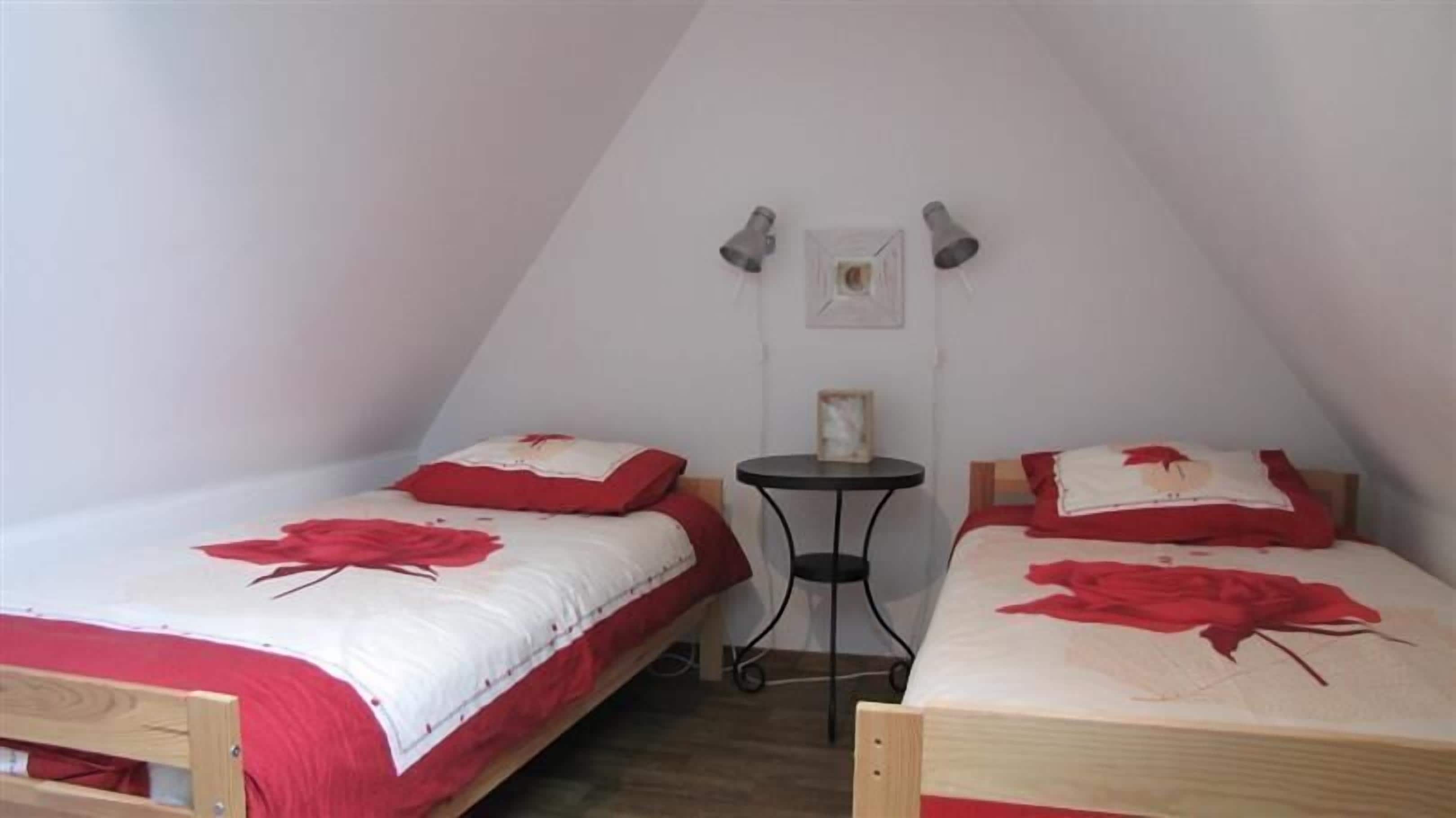 Un dormitorio estilo altillo pequeño y triangular con dos camas, cada una con ropa de cama y almohadas de estampado floral rojo. Hay una mesita redonda pequeña y dos lámparas fijadas a la pared.