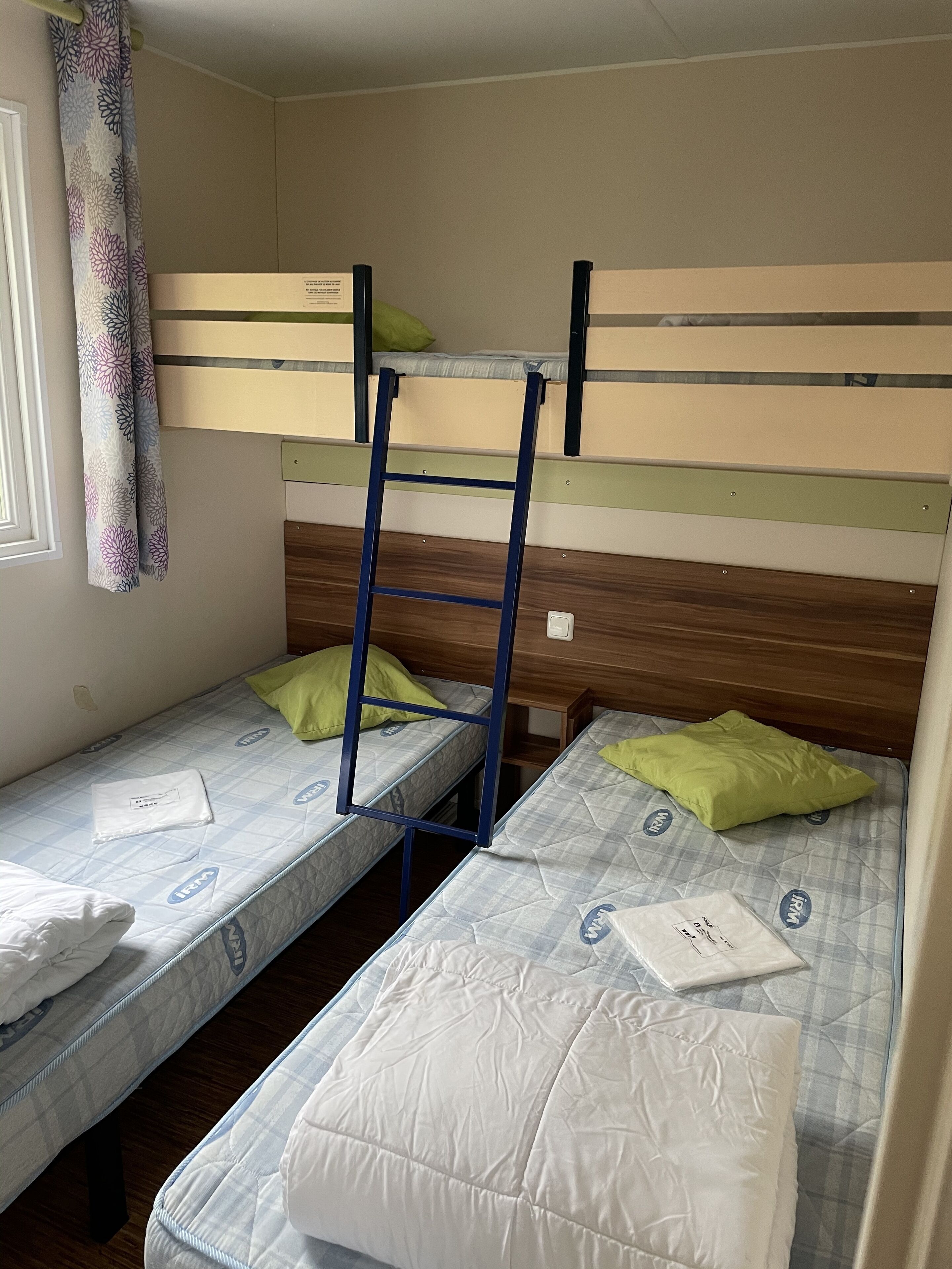 A dormitory room with bunk beds, a ladder, and a window with a curtain.