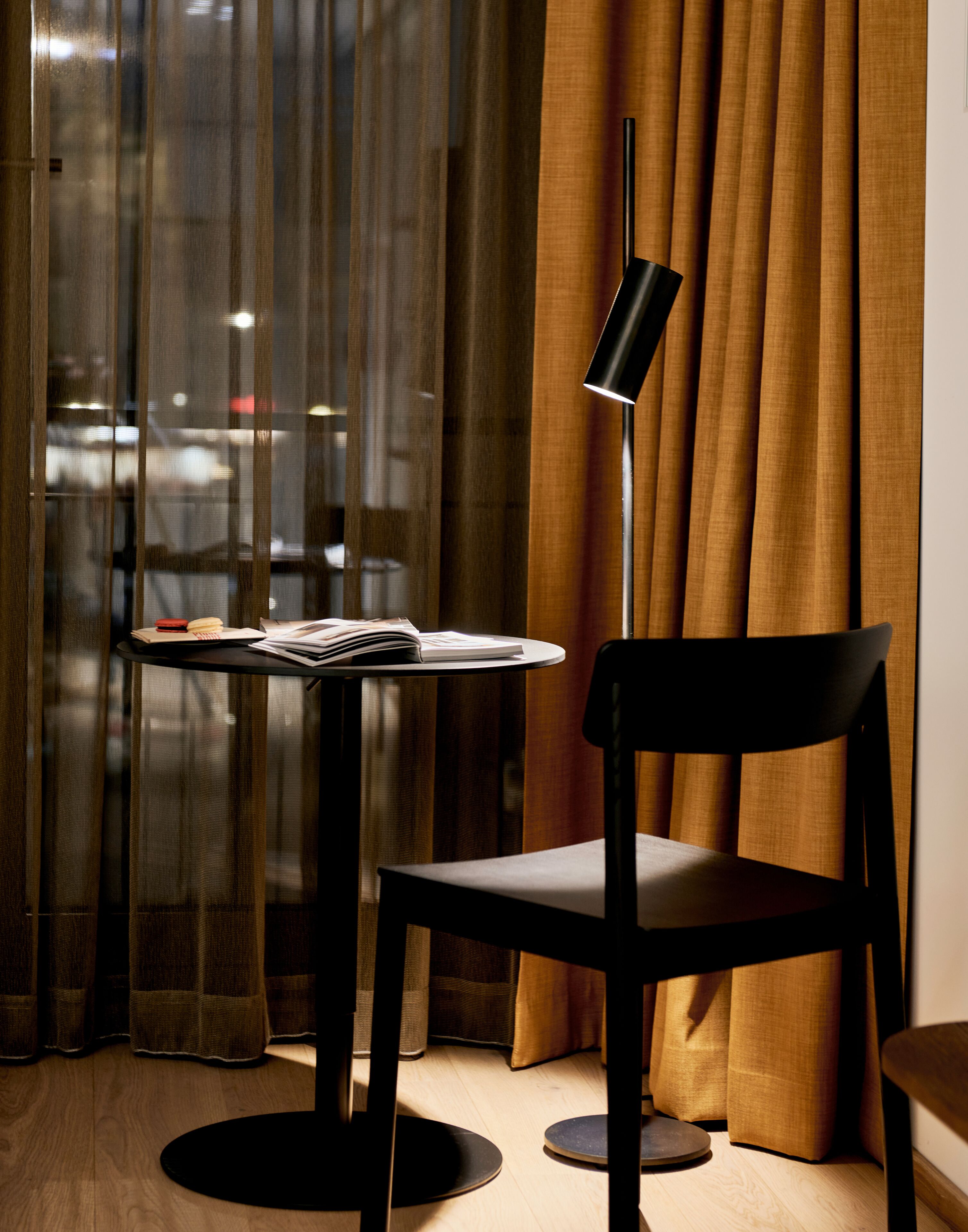 Una habitación moderna con una mesa redonda, una silla negra, una lámpara de pie y vistas a través de una ventana con cortinas.