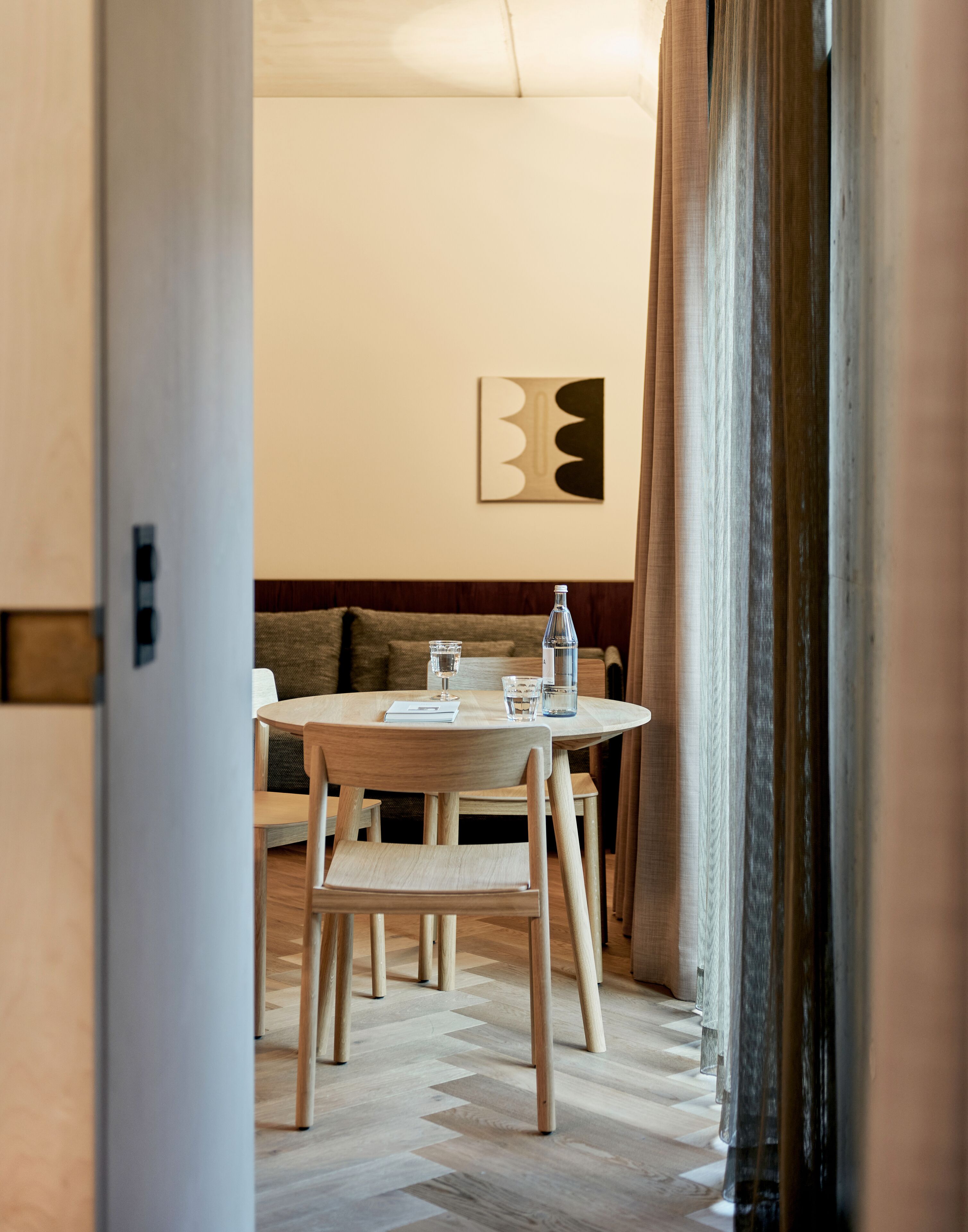 Un comedor moderno con una mesa redonda, sillas de madera y un sofá. Hay una botella y copas sobre la mesa, y una obra de arte en la pared.