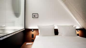 Una habitación de hotel moderna con una cama grande, una mesita de noche pequeña, un cuadro enmarcado en la pared y una ventana con persianas.