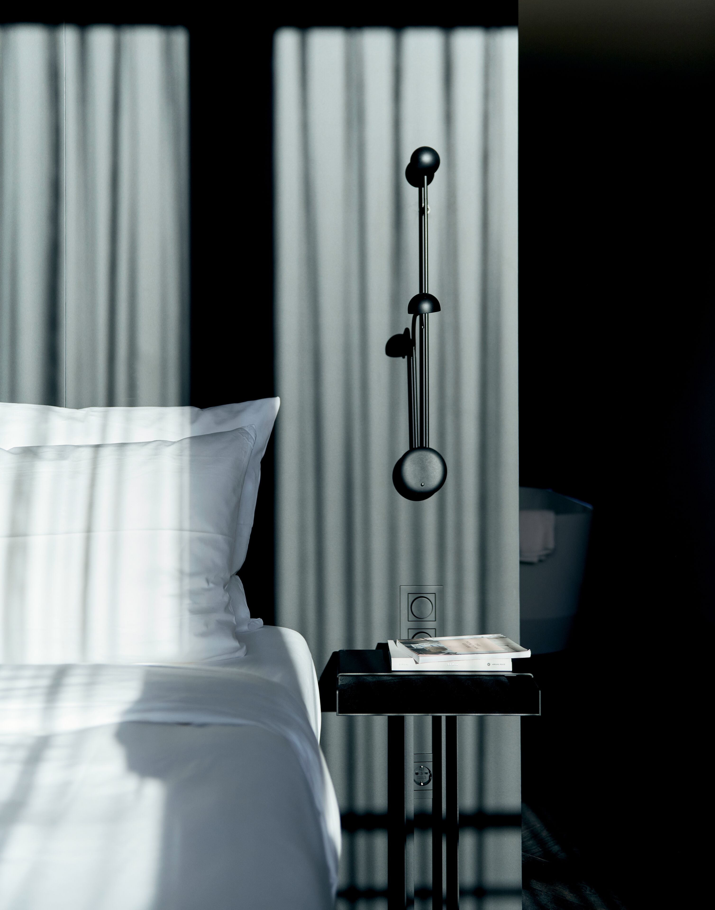 A neatly made bed with white linens, a bedside table with a lamp, and a window with vertical blinds.