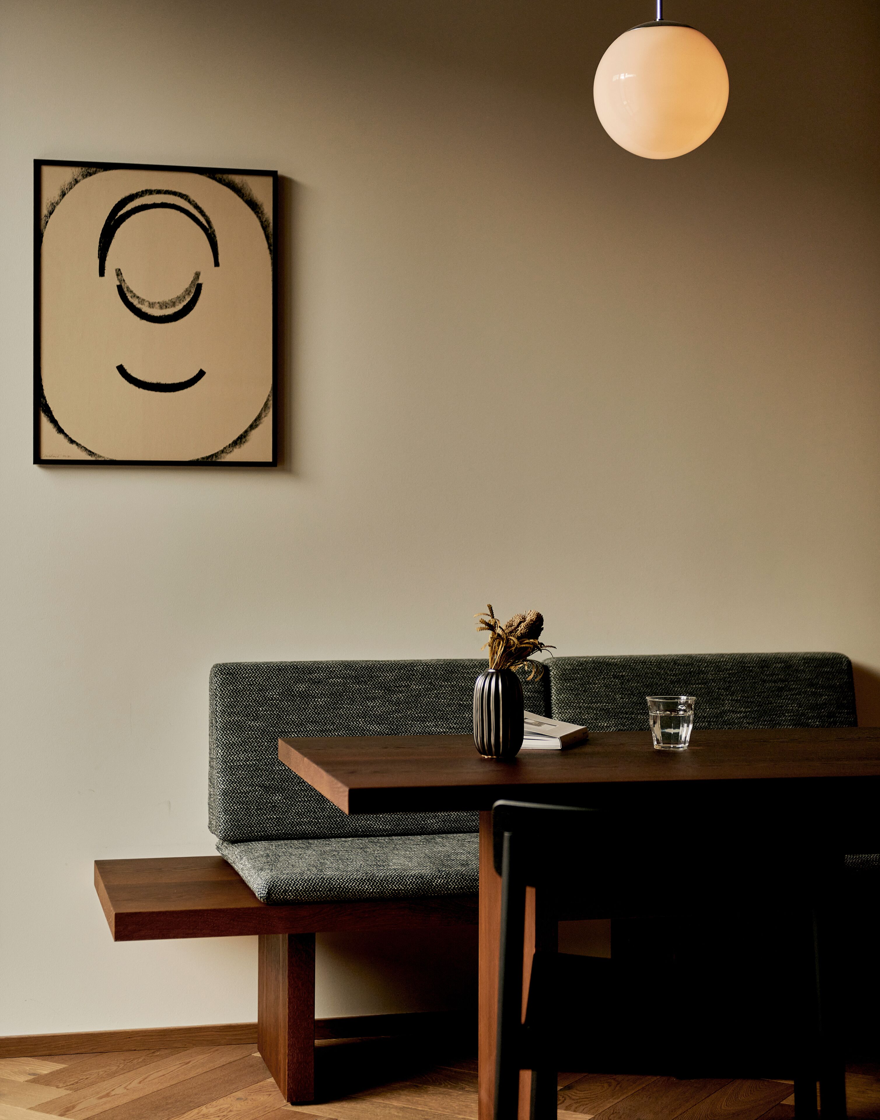A dining area with a wooden table, a bench, a vase with dried flowers, and a glass of water.