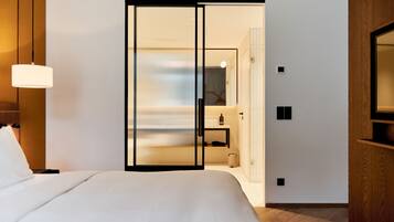 Habitación de hotel moderna con una cama grande, una puerta corredera de vidrio que da a un baño y suelo de madera.