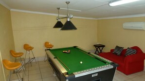 Una mesa de billar con cuatro taburetes, un sofá rojo y un ventilador de techo en una habitación con paredes amarillas y suelo de baldosas.