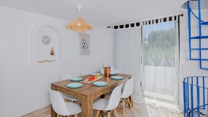 A dining area with a wooden table, white chairs, a hanging light fixture, and a window with sheer curtains. - Eco Beachfront Villa (Gennadi)