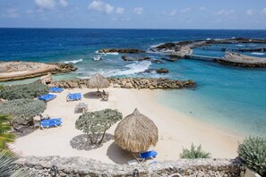 Un complexe hôtelier de bord de mer doté de parasols en chaume, de chaises longues bleues et d’une côte rocheuse.
