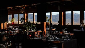 Un restaurante tenuemente iluminado con mesas dispuestas para comer, ventanas amplias y plantas en macetas.