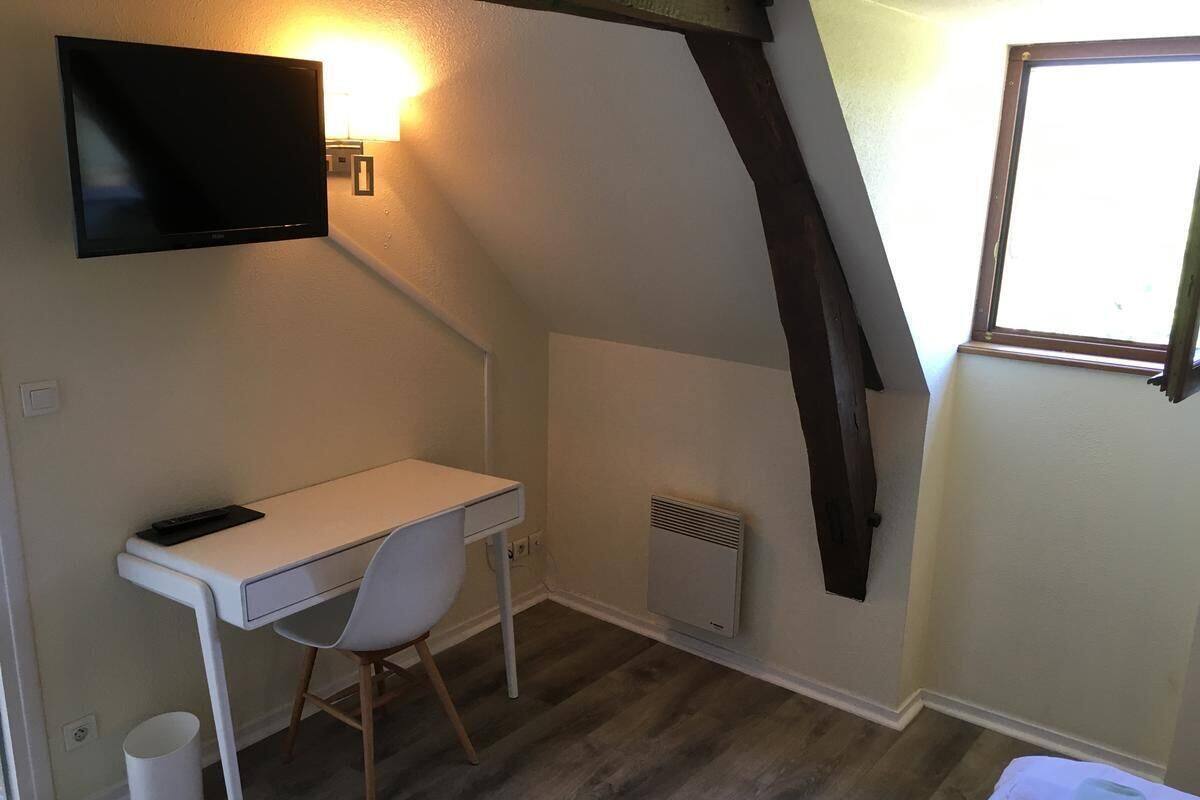 Una habitación pequeña y funcional con un escritorio blanco, una silla, un televisor montado, un radiador y una ventana.
