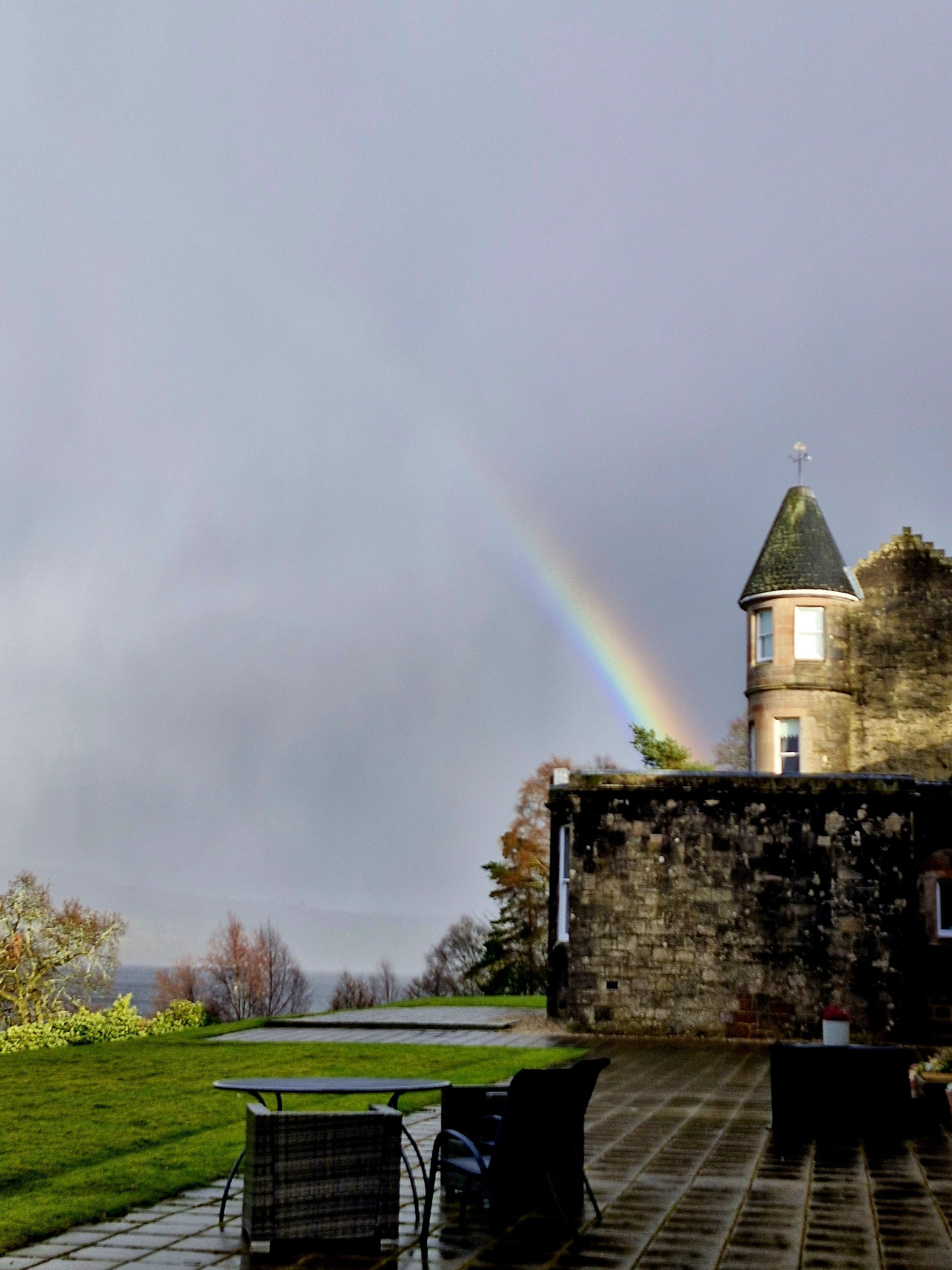 A stone building with a tower, a grassy area with a table and chairs, and a rainbow in the sky.
