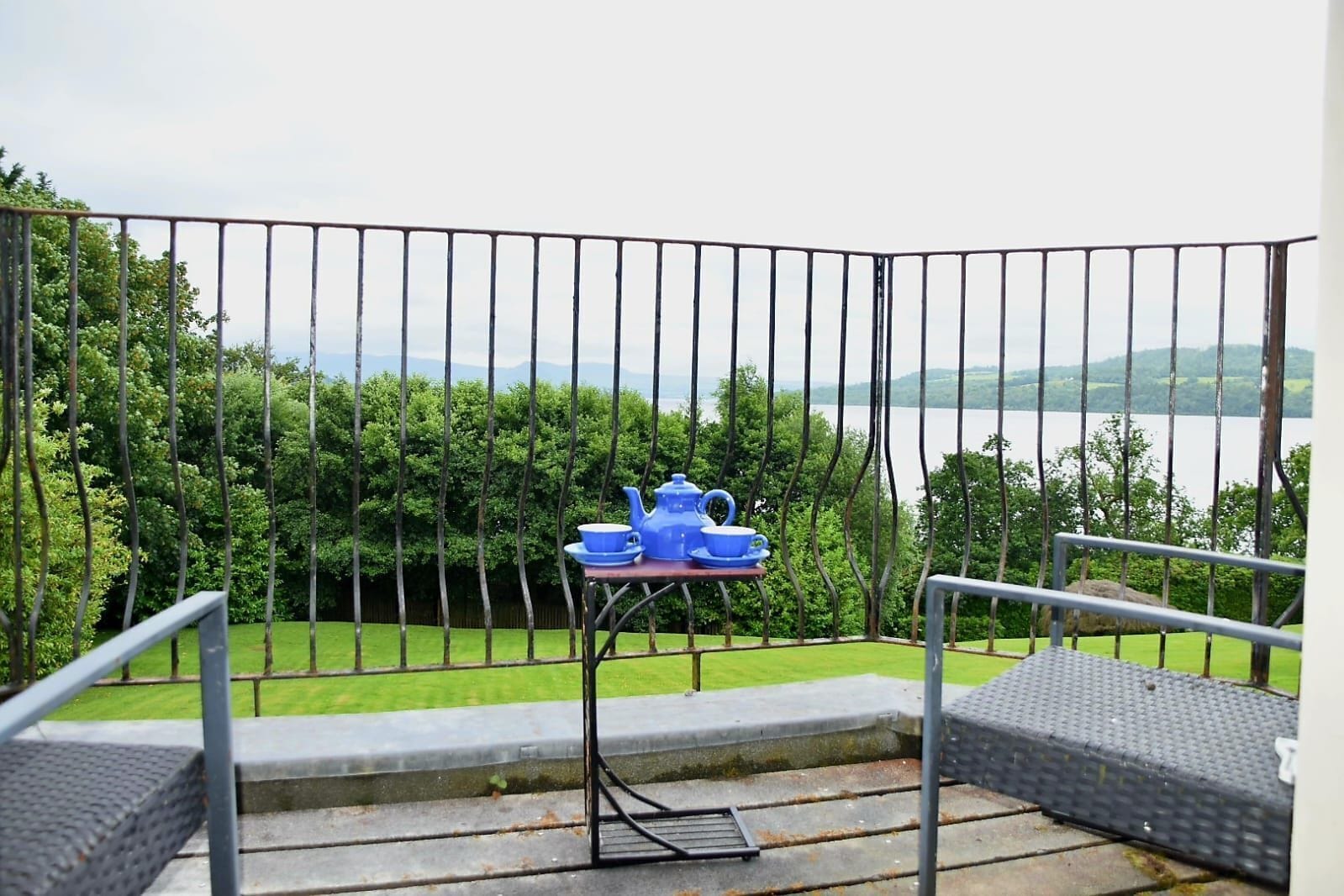 Un balcón con barandilla de metal, una mesita con una tetera azul y tazas, y vistas a un lago y follaje.