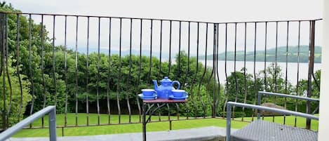 Un balcón con barandilla de metal, una mesita con una tetera azul y tazas, y vistas a un lago y follaje.