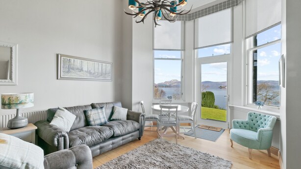 A modern living room with a grey sofa, a round table, and a view of a lake through large windows. - No 3 Lomond Castle - Sleeps 4 - sleeps 4 guests  in 2 bedrooms (ALEXANDRIA)