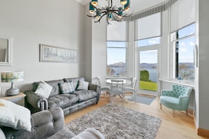 A modern living room with a grey sofa, a round table, and a view of a lake through large windows.