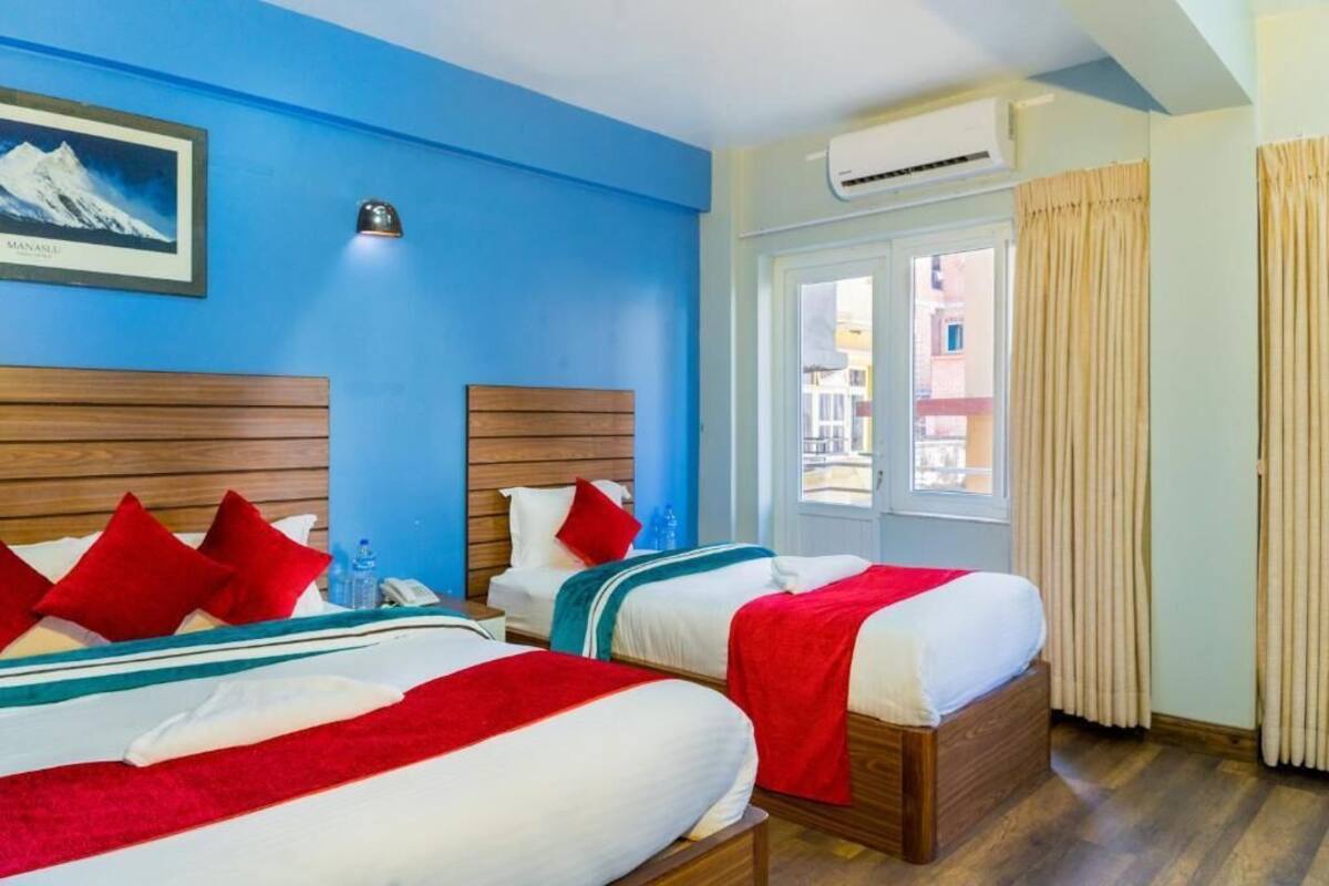 Une chambre d’hôtel avec deux lits, des têtes de lit en bois, une literie rouge et blanche, une fenêtre avec des rideaux et un appareil de climatisation.