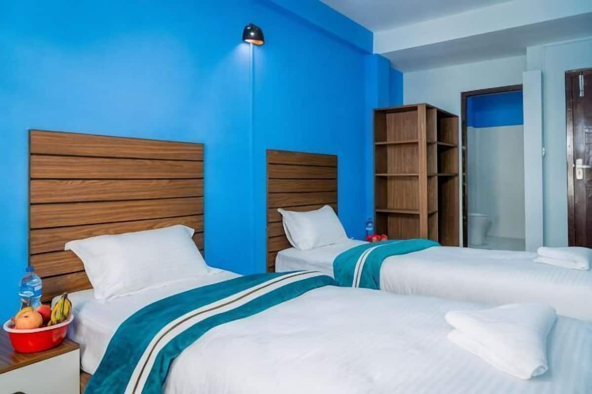 Quarto de hotel com duas camas, cabeceiras de madeira, parede azul, mesa de cabeceira com uma garrafa de água e uma tigela vermelha.