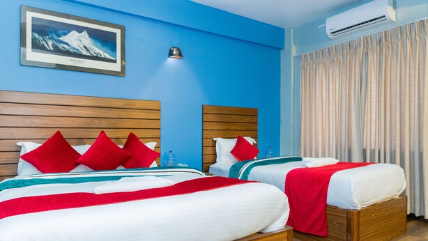 A hotel room with two beds, wooden headboards, red and white bedding, a framed picture of a mountain, an air conditioning unit, and a window with curtains. - Hotel Nepal Bhumi (Kathmandu)