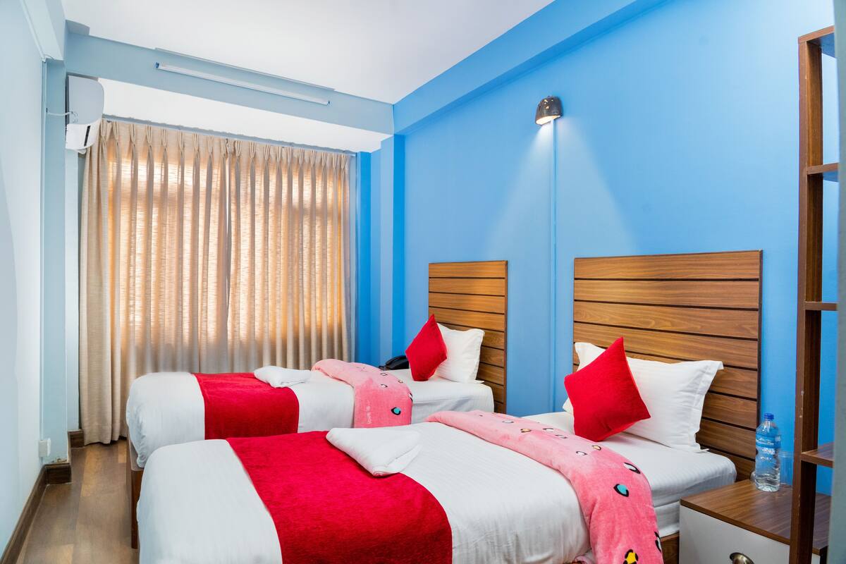 Une chambre d’hôtel avec deux lits, chacun doté de draps rouges et blancs, d’une tête de lit en bois, d’une table de chevet avec une bouteille d’eau et d’une fenêtre avec des rideaux.