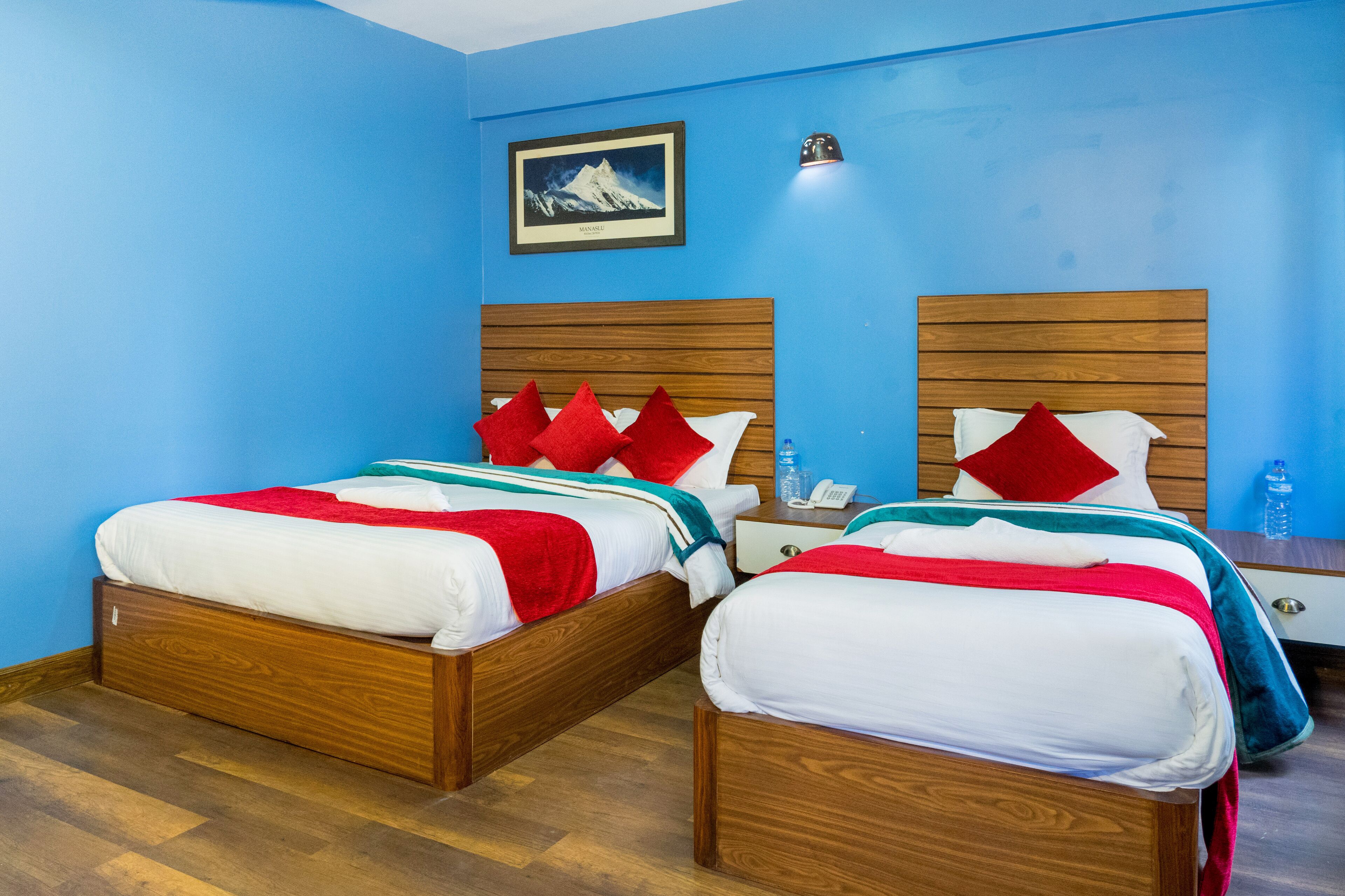 A hotel room with two wooden beds, red and white bedding, a framed picture on the wall, and a bedside table with a phone and water bottle.
