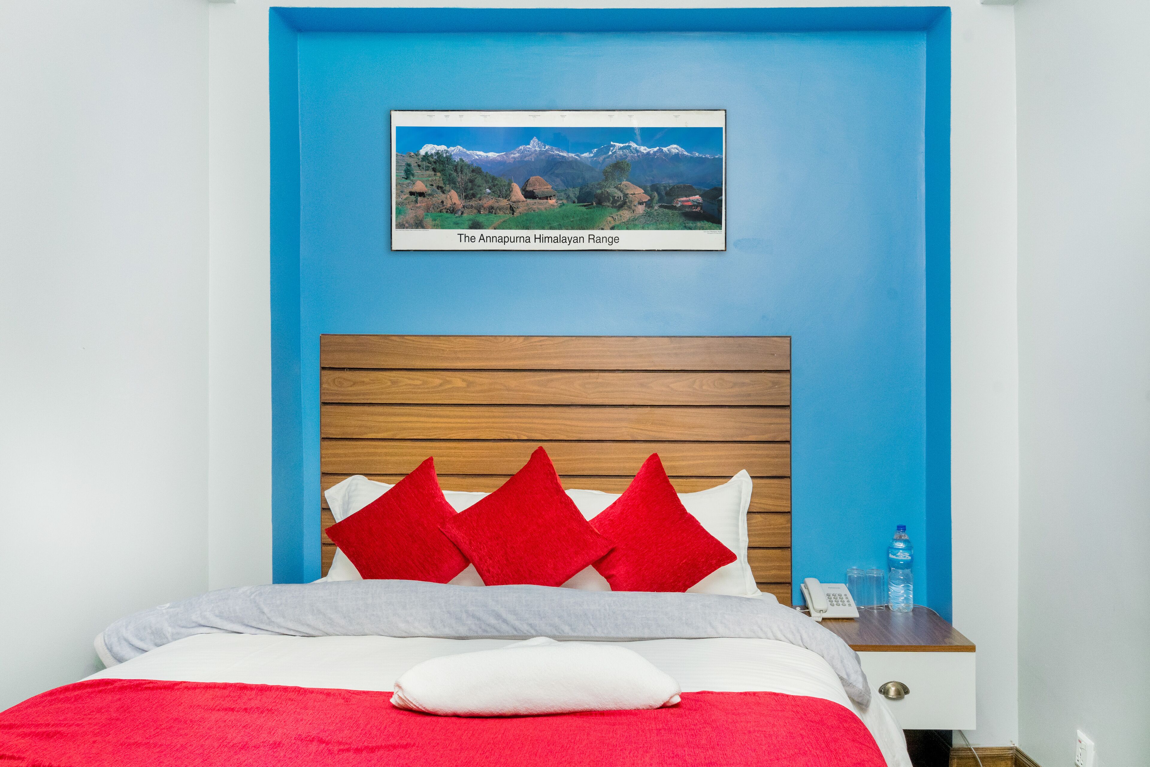 A bedroom with a blue wall, a wooden headboard, a bed with red and white bedding, and a framed picture of the Annapurna Himalayan Range.