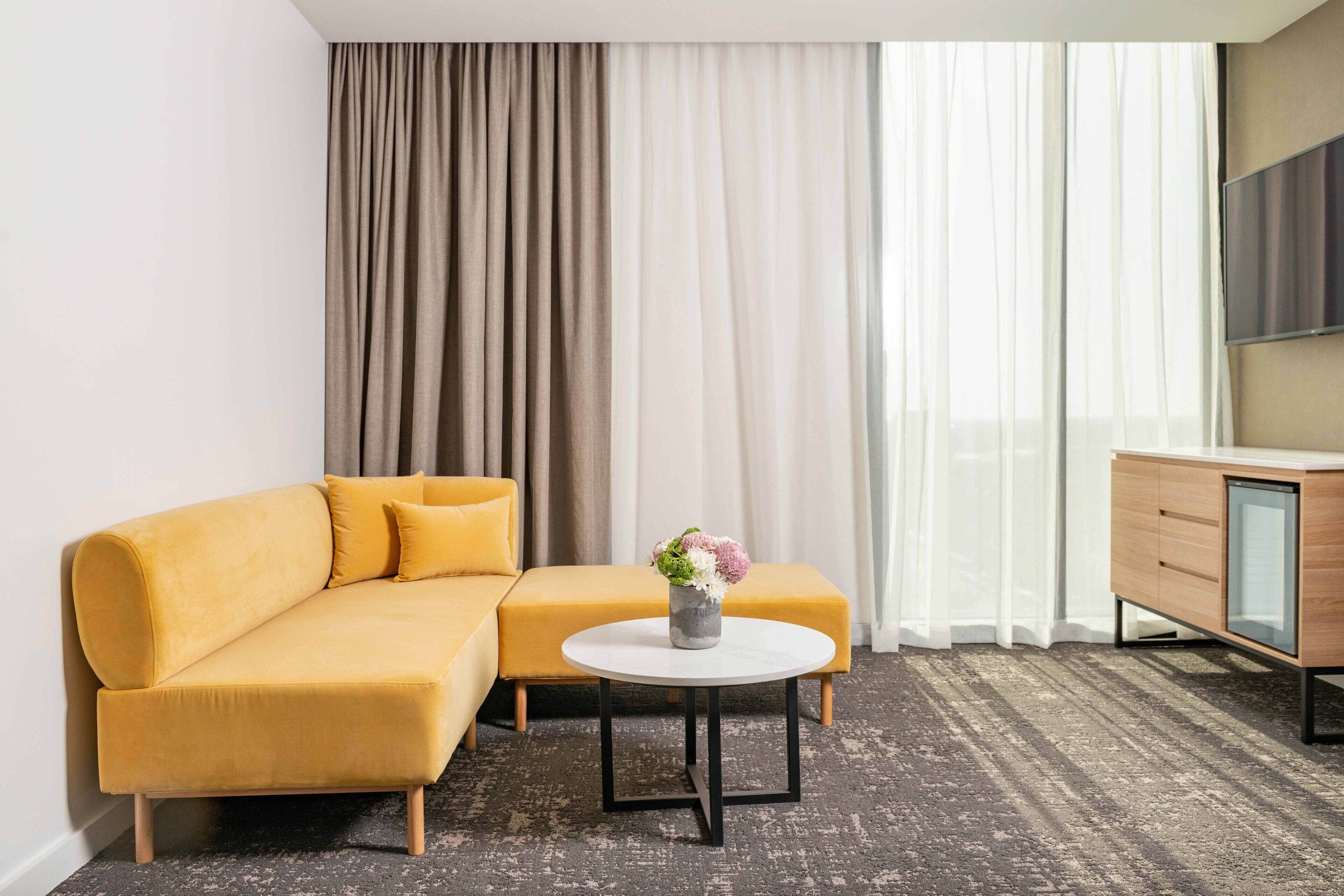 A modern hotel room with a yellow sofa, a round coffee table with a vase of flowers, a wooden cabinet, and a large window with sheer curtains.