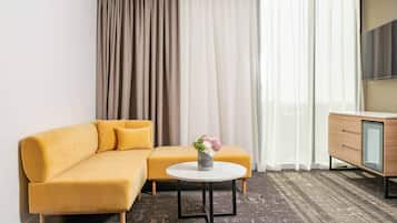 A modern hotel room with a yellow sofa, a round coffee table with a vase of flowers, a wooden cabinet, and a large window with sheer curtains.