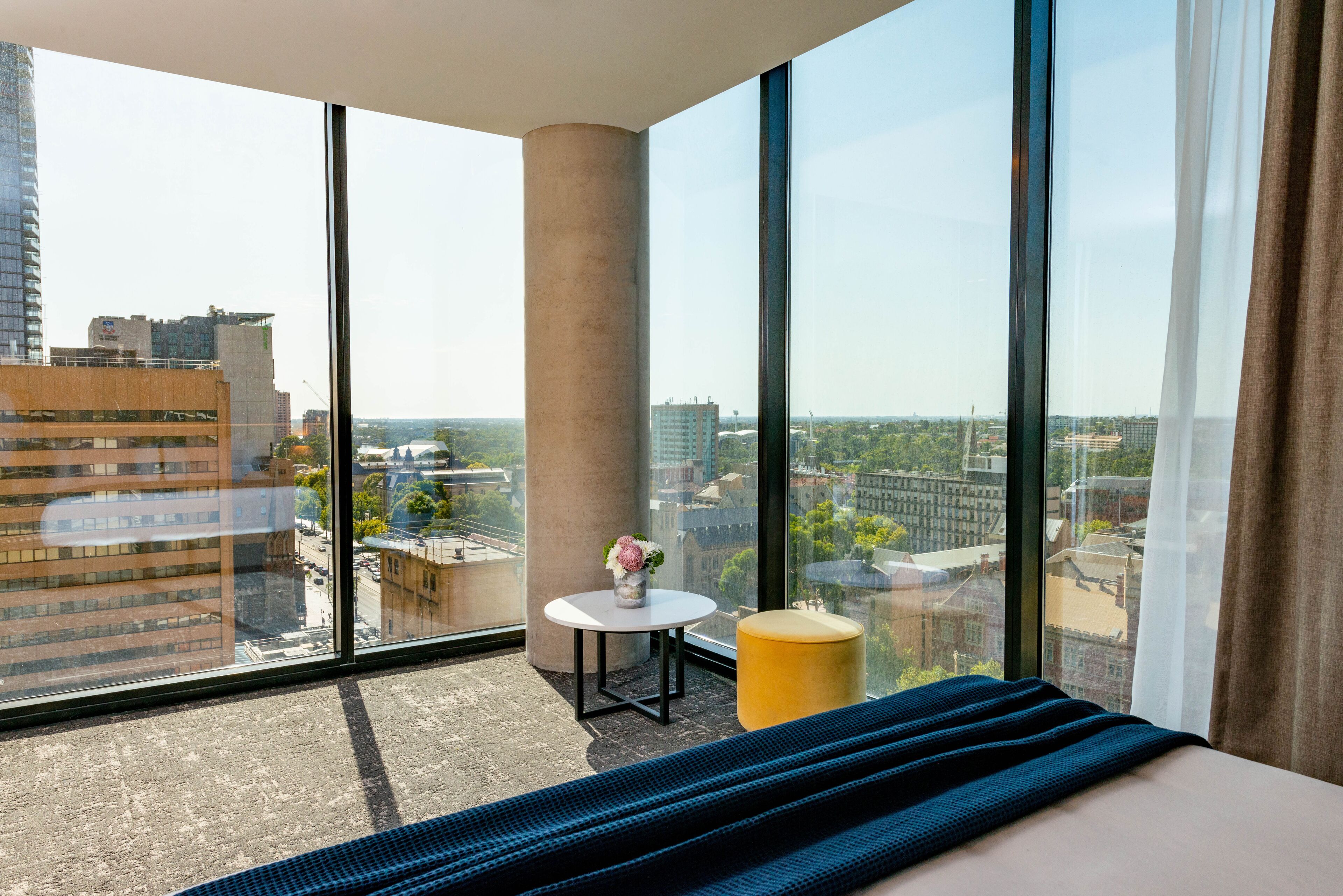 A modern hotel room with large windows offering a city view, a round table with a vase, and a yellow ottoman.