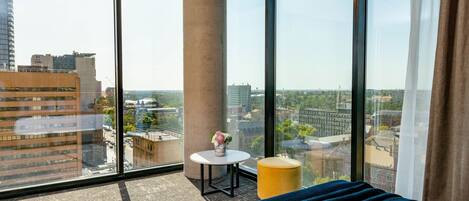 A modern hotel room with large windows offering a city view, a round table with a vase, and a yellow ottoman.