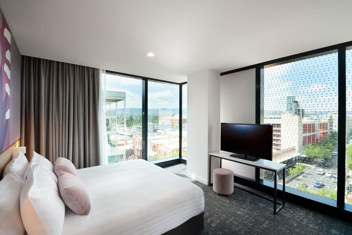 Une chambre d’hôtel moderne dotée d’un grand lit, d’un bureau avec une télévision et offrant une vue sur la ville grâce à de grandes fenêtres.