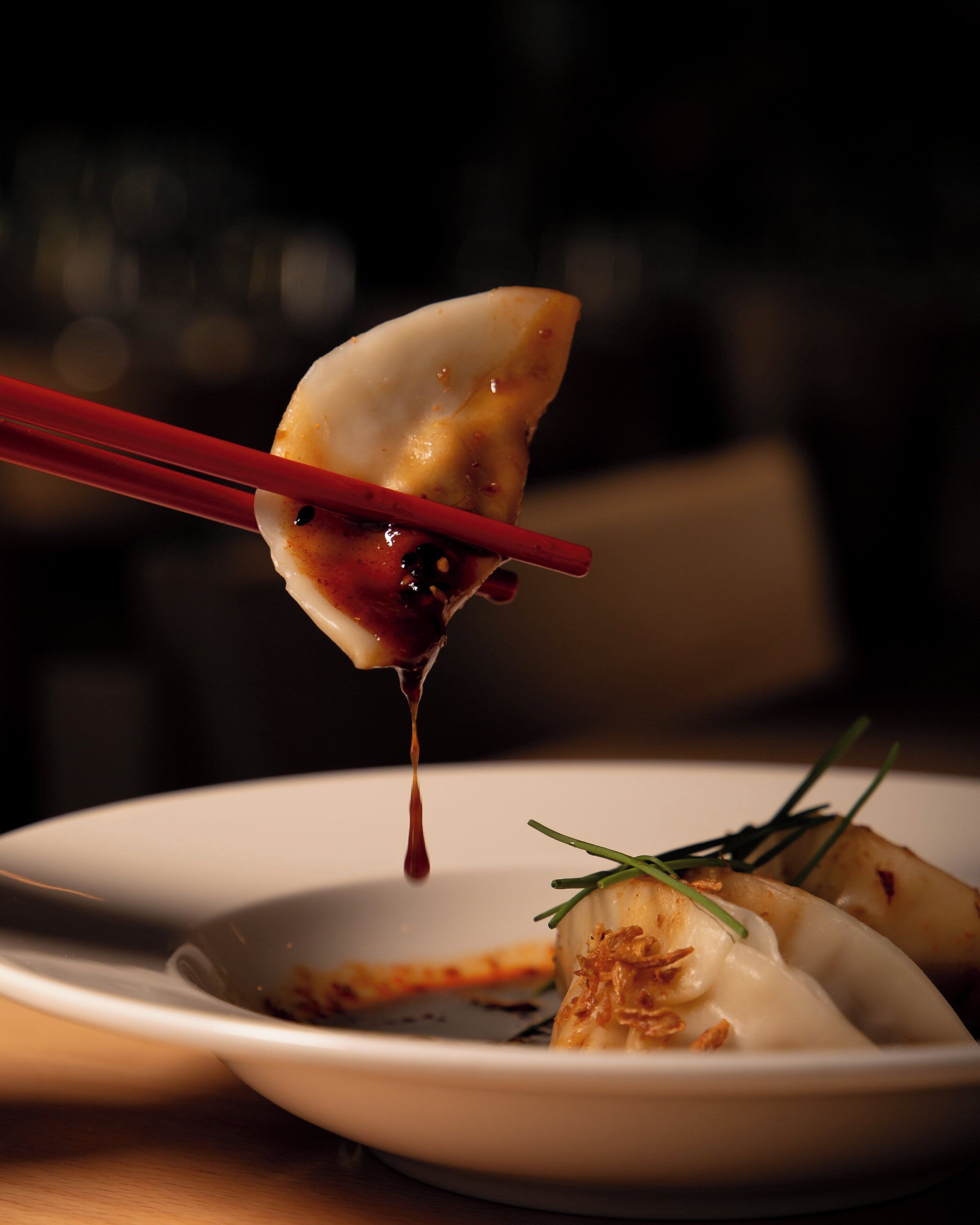 Une assiette contenant des raviolis et une sauce, le tout soulevé à l’aide de baguettes.