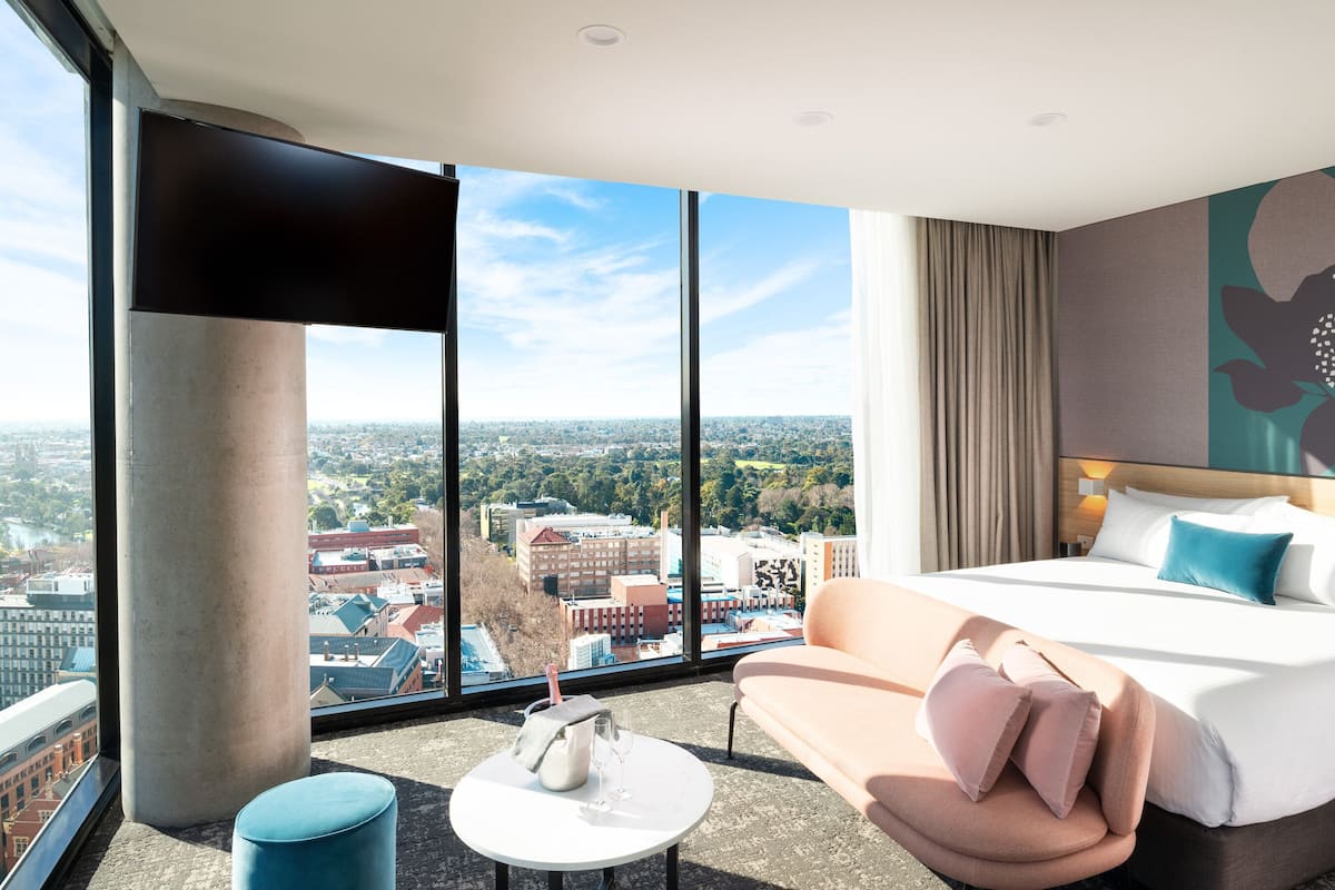 Une chambre d’hôtel moderne dotée d’une grande fenêtre donnant sur la ville, d’un lit avec des oreillers, d’une petite table ronde avec une bouteille et des verres, et d’une chaise bleue.