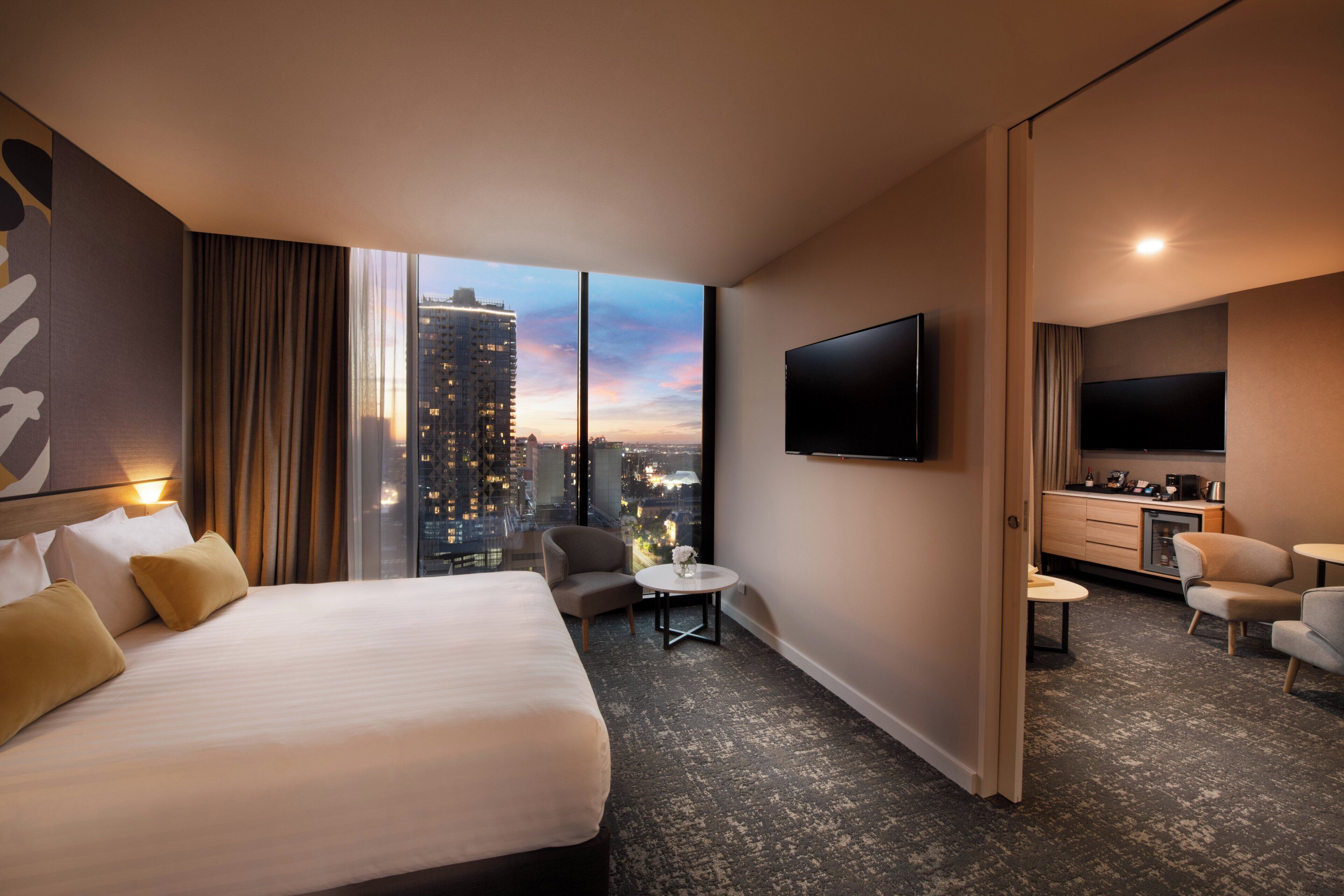 A hotel room with a large bed, a TV, a small table, a chair, and a view of a cityscape.