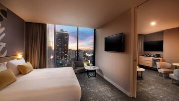 A hotel room with a large bed, a TV, a small table, a chair, and a view of a cityscape.