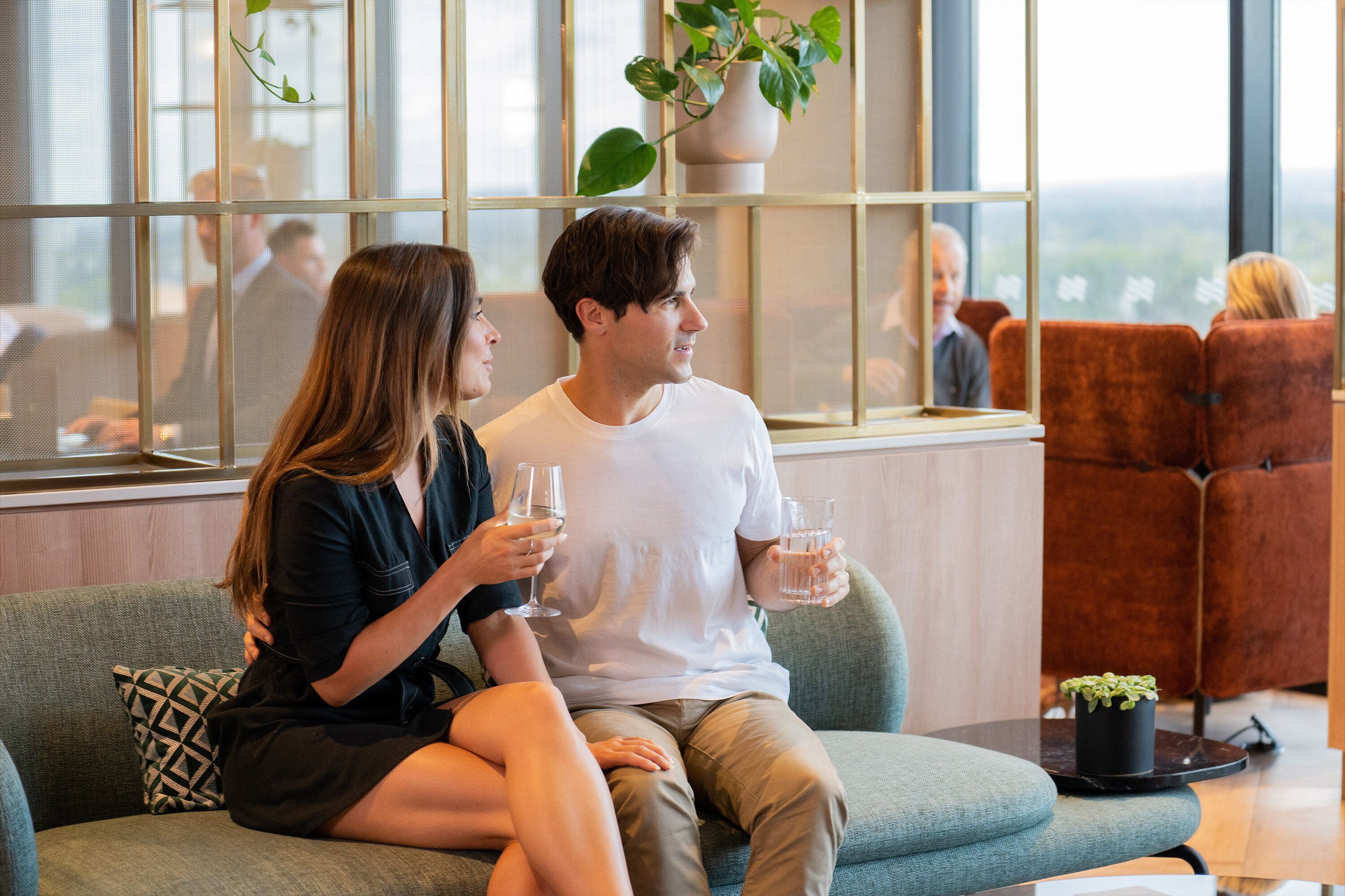 A couple sitting on a sofa in a modern lounge area with large windows, a small table, and a potted plant.