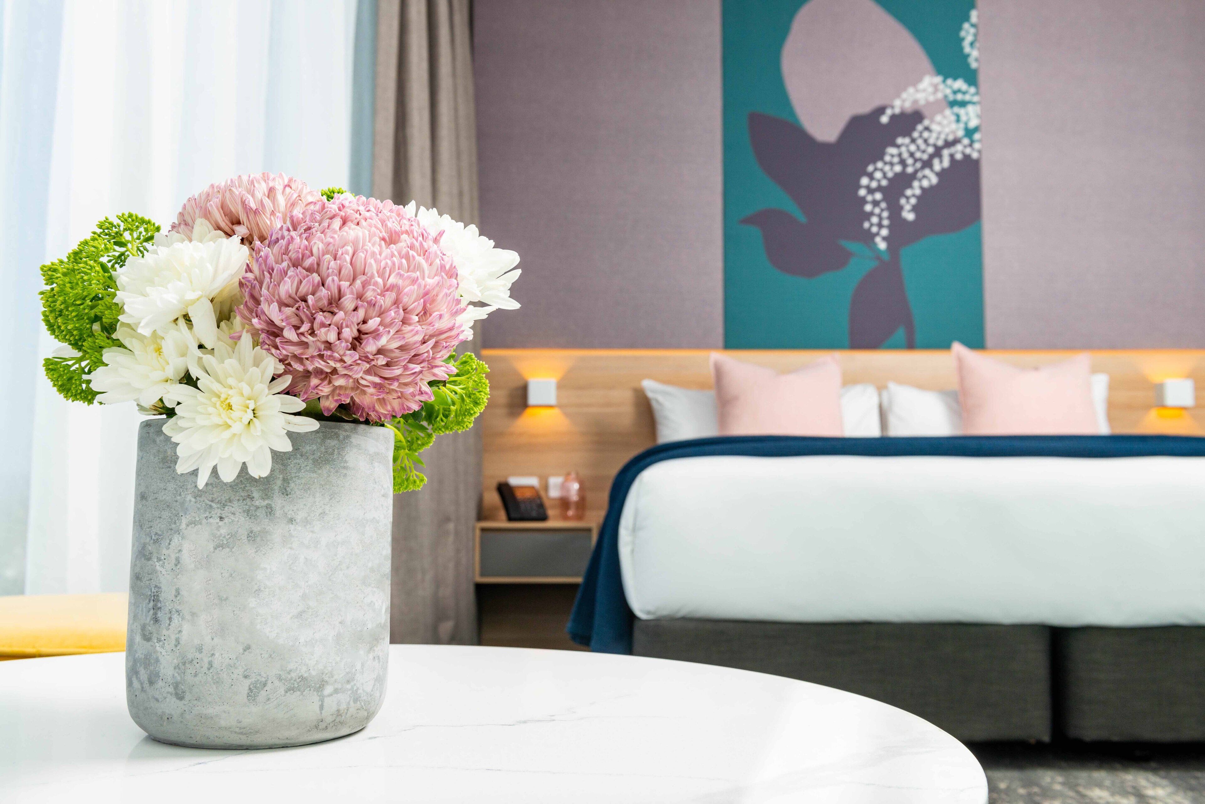 A hotel room with a bed, a nightstand, and a vase of flowers on a round table.