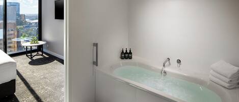 A modern bathroom with a bathtub, a small table with bottles, and a view of the city through large windows.