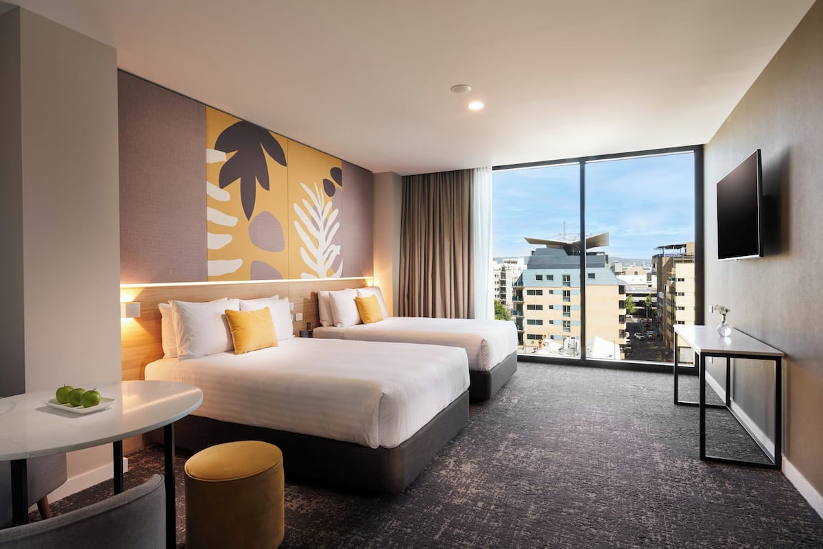 Une chambre d’hôtel avec deux lits, une petite table avec des pommes, une télévision et une grande fenêtre donnant sur la ville.